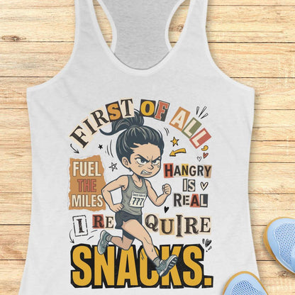 white racerback tank top featuring a cartoon runner wearing a race bib and running shoes, surrounded by bold typography. The design reads: “First of all, fuel the miles. Hangry is real. I require snacks.” The artwork uses a playful mix of fonts, yellow, black, beige, and coral accents, with doodle-style arrows, stars, and hearts. The tank top is displayed on a light wood surface with running shoes partially visible in the corners.