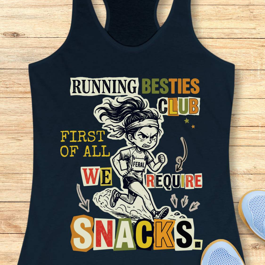 A navy blue racerback tank top with a colorful graphic print of a cartoon runner and the text 'RUNNING BESTIES CLUB FIRST OF ALL WE REQUIRE SNACKS.', displayed flat on a light wood background.