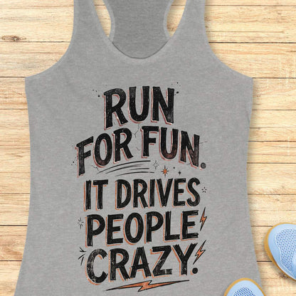 grey racerback tank top featuring a bold typographic running design. The text reads: “Run for fun. It drives people crazy.” in distressed black lettering with vintage-style shading. Decorative elements include stars, sparkles, curved accent lines, and lightning bolts. The tank top is displayed flat on a light wood background with partial blue running shoes visible in the lower corners.