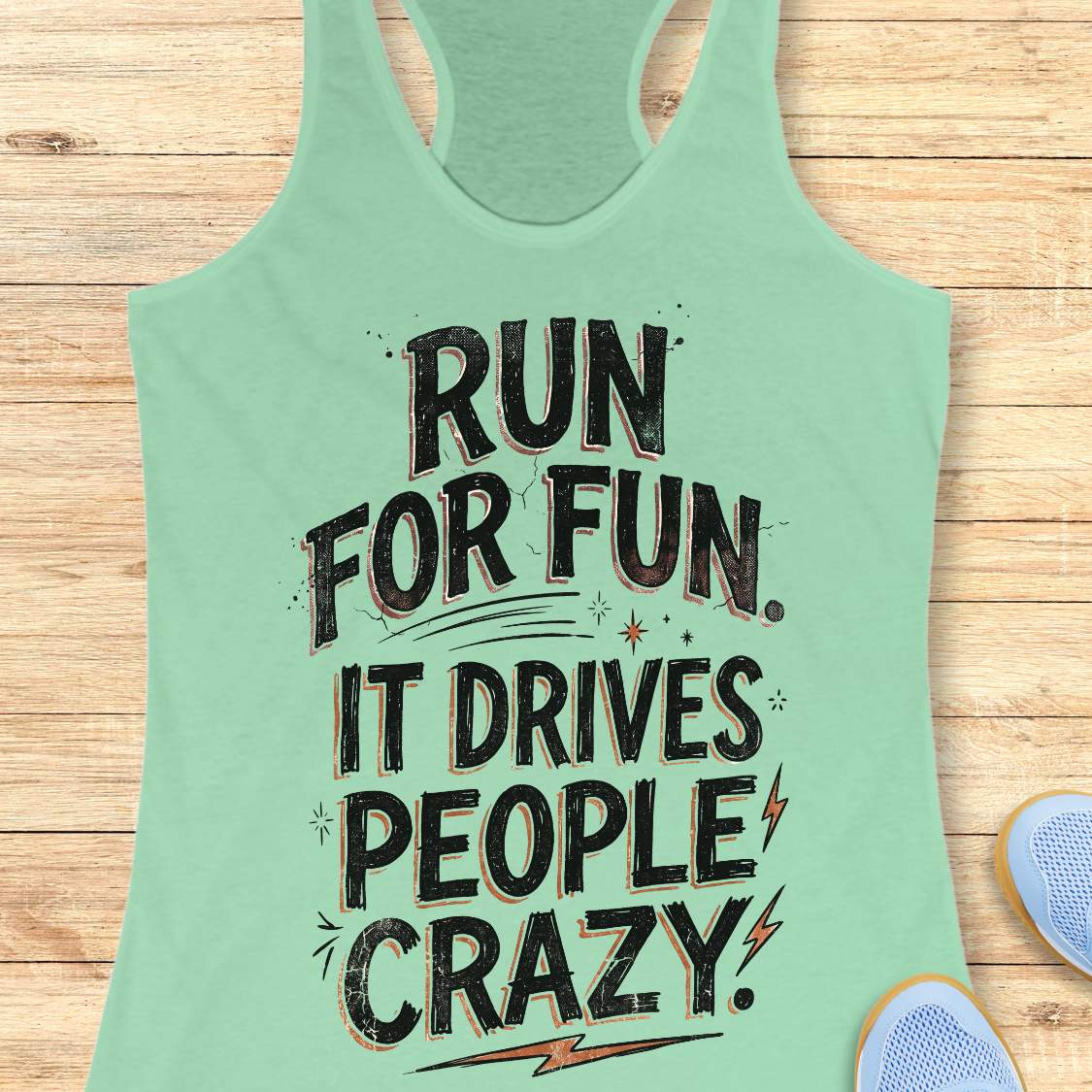 Mint green racerback tank top featuring a bold typographic running design. The text reads: “Run for fun. It drives people crazy.” in distressed black lettering with vintage-style shading. Decorative elements include stars, sparkles, curved accent lines, and lightning bolts. The tank top is displayed flat on a light wood background with partial blue running shoes visible in the lower corners.