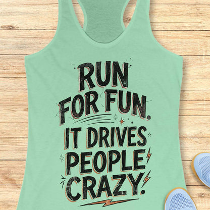 Mint green racerback tank top featuring a bold typographic running design. The text reads: “Run for fun. It drives people crazy.” in distressed black lettering with vintage-style shading. Decorative elements include stars, sparkles, curved accent lines, and lightning bolts. The tank top is displayed flat on a light wood background with partial blue running shoes visible in the lower corners.