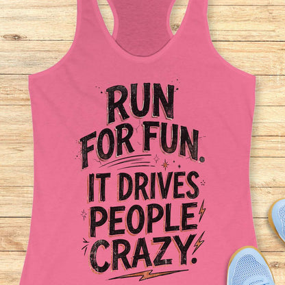 pink racerback tank top featuring a bold typographic running design. The text reads: “Run for fun. It drives people crazy.” in distressed black lettering with vintage-style shading. Decorative elements include stars, sparkles, curved accent lines, and lightning bolts. The tank top is displayed flat on a light wood background with partial blue running shoes visible in the lower corners.