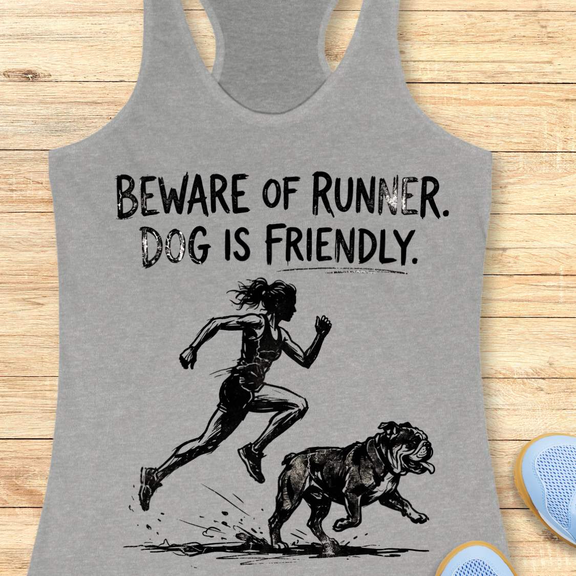 grey racerback tank top featuring the humorous slogan, “BEWARE OF RUNNER. DOG IS FRIENDLY.” in distressed black lettering above a black sketch-style illustration of a runner sprinting behind a friendly bulldog. The design playfully suggests the runner is more intimidating than the dog, making it a fun graphic for dog-loving runners.