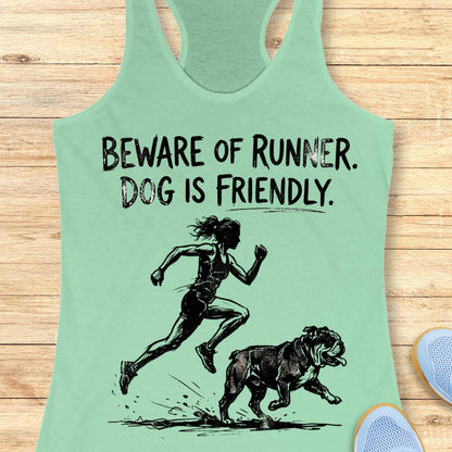 mint racerback tank top featuring the humorous slogan, “BEWARE OF RUNNER. DOG IS FRIENDLY.” in distressed black lettering above a black sketch-style illustration of a runner sprinting behind a friendly bulldog. The design playfully suggests the runner is more intimidating than the dog, making it a fun graphic for dog-loving runners.