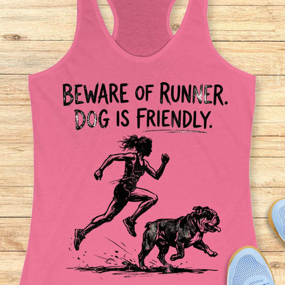 pink racerback tank top featuring the humorous slogan, “BEWARE OF RUNNER. DOG IS FRIENDLY.” in distressed black lettering above a black sketch-style illustration of a runner sprinting behind a friendly bulldog. The design playfully suggests the runner is more intimidating than the dog, making it a fun graphic for dog-loving runners.