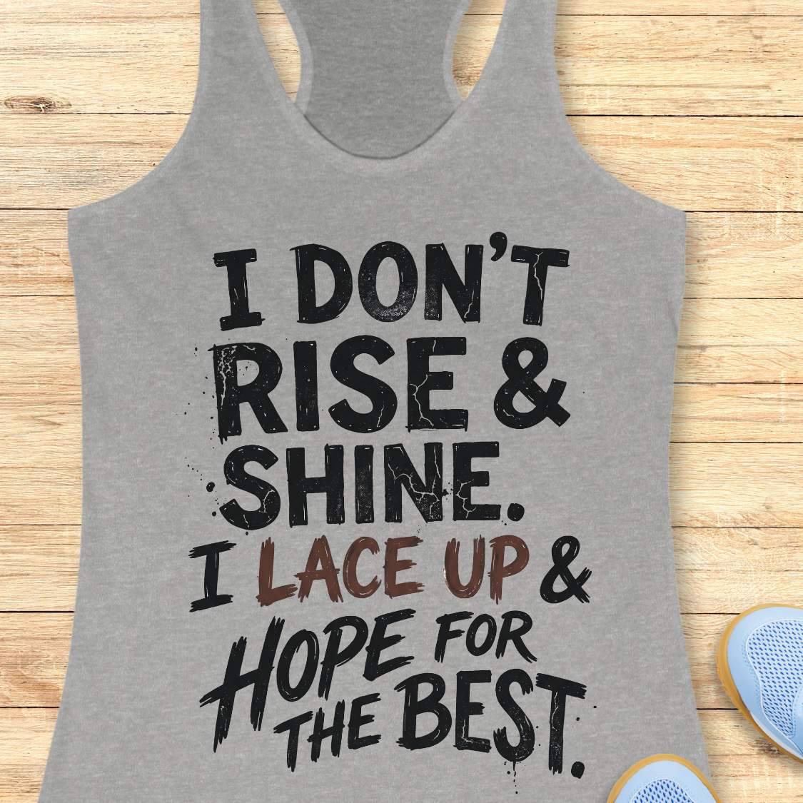 grey racerback tank top featuring a bold, distressed typography design that reads: “I DON’T RISE & SHINE. I LACE UP & HOPE FOR THE BEST.” The text is displayed in a mix of black and brown brush-style lettering, creating a playful and relatable message for runners who prefer determination over morning enthusiasm. The humorous running-themed design is ideal for athletes, joggers, and fitness enthusiasts.