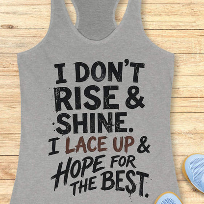 grey racerback tank top featuring a bold, distressed typography design that reads: “I DON’T RISE & SHINE. I LACE UP & HOPE FOR THE BEST.” The text is displayed in a mix of black and brown brush-style lettering, creating a playful and relatable message for runners who prefer determination over morning enthusiasm. The humorous running-themed design is ideal for athletes, joggers, and fitness enthusiasts.