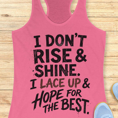 pink racerback tank top featuring a bold, distressed typography design that reads: “I DON’T RISE & SHINE. I LACE UP & HOPE FOR THE BEST.” The text is displayed in a mix of black and brown brush-style lettering, creating a playful and relatable message for runners who prefer determination over morning enthusiasm. The humorous running-themed design is ideal for athletes, joggers, and fitness enthusiasts.