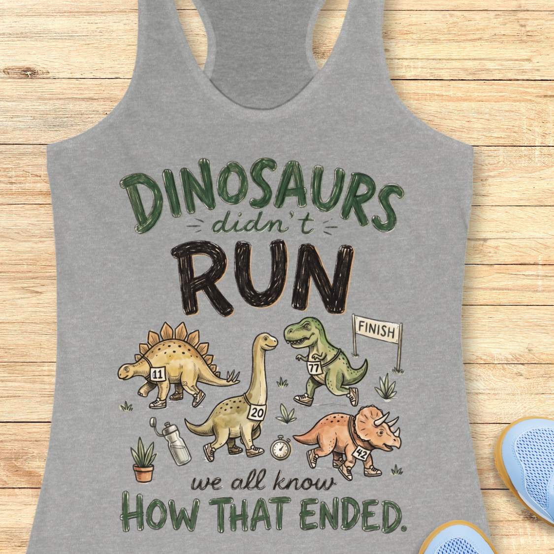 grey racerback tank top featuring the humorous running-themed phrase, “DINOSAURS didn’t RUN we all know HOW THAT ENDED.” The design includes cartoon dinosaurs wearing race bibs and running shoes, jogging toward a finish line. Small race-themed elements such as a water bottle, stopwatch, and plants add detail to the playful illustration. The fun, motivational graphic combines running humor with a lighthearted take on prehistoric extinction, making it ideal for runners and fitness enthusiasts.