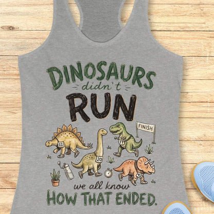 grey racerback tank top featuring the humorous running-themed phrase, “DINOSAURS didn’t RUN we all know HOW THAT ENDED.” The design includes cartoon dinosaurs wearing race bibs and running shoes, jogging toward a finish line. Small race-themed elements such as a water bottle, stopwatch, and plants add detail to the playful illustration. The fun, motivational graphic combines running humor with a lighthearted take on prehistoric extinction, making it ideal for runners and fitness enthusiasts.