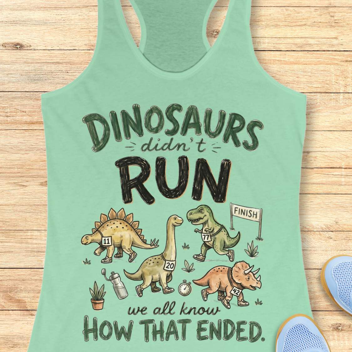 Mint green racerback tank top featuring the humorous running-themed phrase, “DINOSAURS didn’t RUN we all know HOW THAT ENDED.” The design includes cartoon dinosaurs wearing race bibs and running shoes, jogging toward a finish line. Small race-themed elements such as a water bottle, stopwatch, and plants add detail to the playful illustration. The fun, motivational graphic combines running humor with a lighthearted take on prehistoric extinction, making it ideal for runners and fitness enthusiasts.