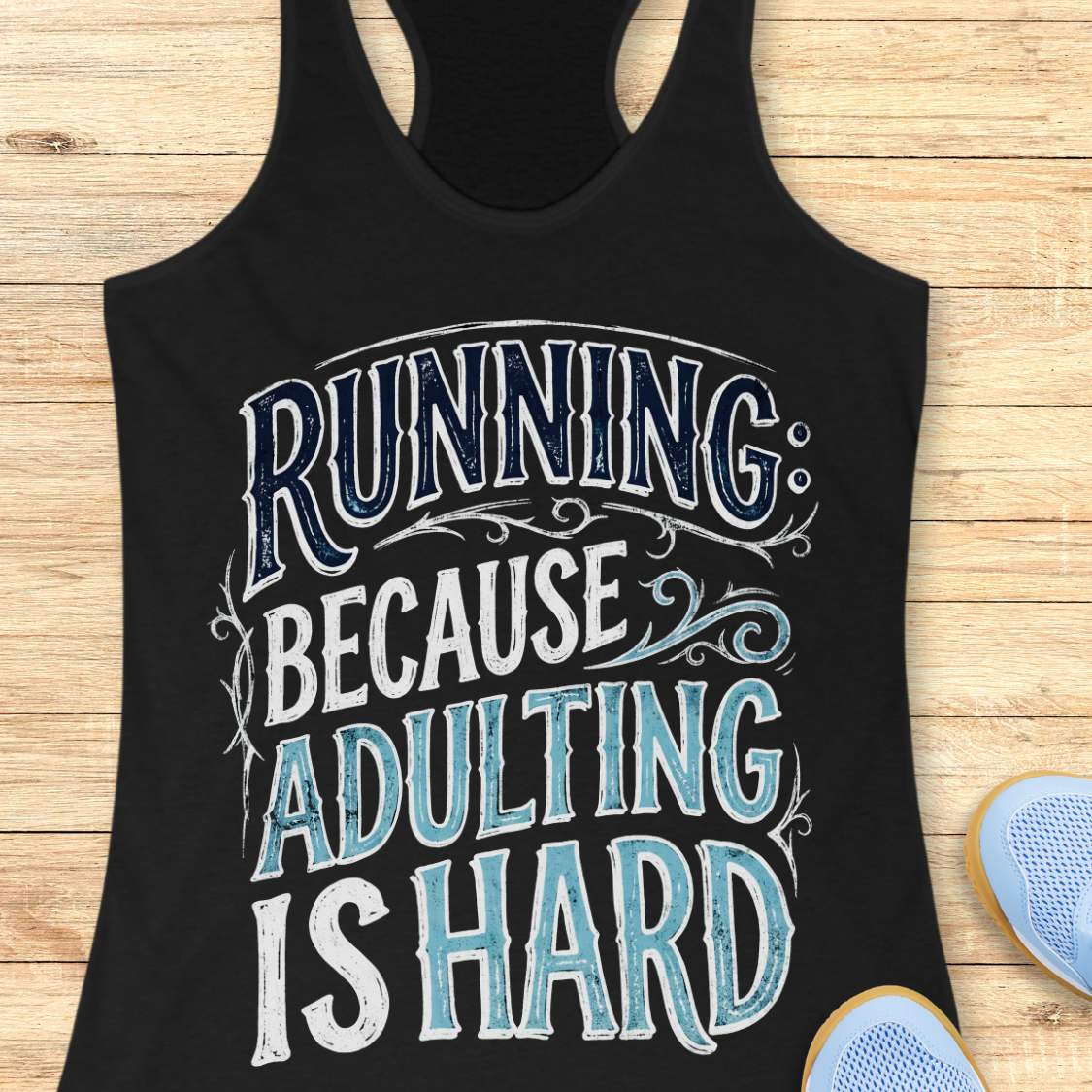 A black racerback tank top laid flat on a light wood background, featuring a large distressed graphic that reads 'RUNNING BECAUSE ADULTING IS HARD' in white and light blue lettering. Partial blue sneakers are visible at the bottom right as styling props.