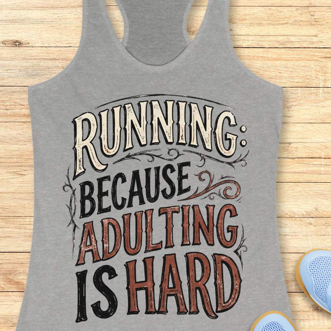 A heather gray racerback tank top with a graphic text print reading 'RUNNING: BECAUSE ADULTING IS HARD' in cream, black, and distressed brown lettering, displayed flat on a light wood plank background.