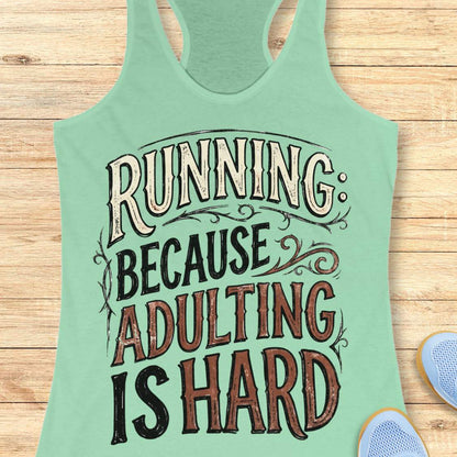 A mint green racerback tank top with a humorous graphic reading 'RUNNING: BECAUSE ADULTING IS HARD' in stylized cream, black, and brown lettering, laid flat on a light wood plank background with partial blue sneakers visible at the bottom right.
