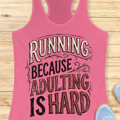 A pink racerback tank top laid flat on a light wood background, featuring the text 'RUNNING BECAUSE ADULTING IS HARD' in stylized cream, black, and brown lettering. Partial blue sneakers are visible at the edges as styling props.