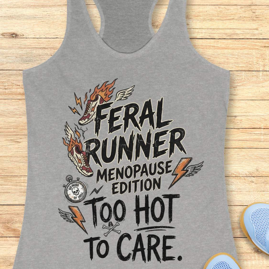 A heather gray racerback tank top with a graphic print reading 'FERAL RUNNER MENOPAUSE EDITION TOO HOT TO CARE.' in black, with orange flames, lightning bolts, a pocket watch, and crossed bones, shown on a wood plank background.