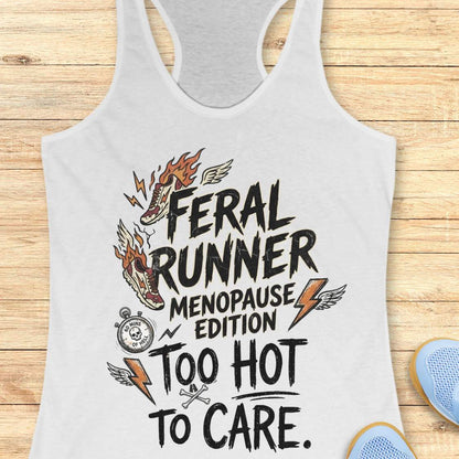 A white racerback tank top laid flat on a light wood background, featuring a graphic print with the text 'FERAL RUNNER MENOPAUSE EDITION TOO HOT TO CARE' and illustrated flames, lightning bolts, wings, and a stopwatch.