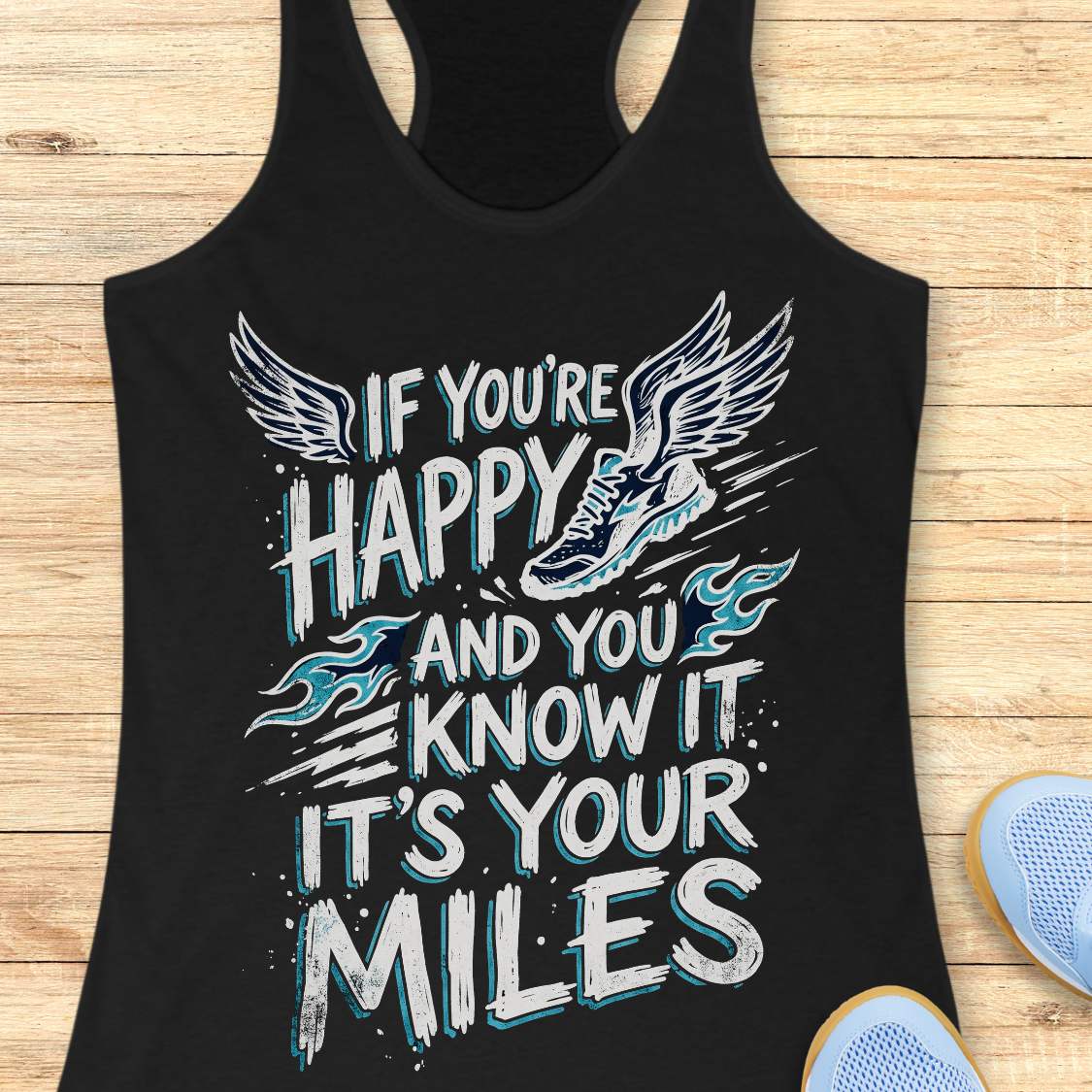 A black racerback tank top laid flat on a light wood background, featuring a large white and light blue graphic with wings, a running shoe, and the text 'IF YOU'RE HAPPY AND YOU KNOW IT IT'S YOUR MILES.' Partial blue shoes are visible at the bottom right as styling props.