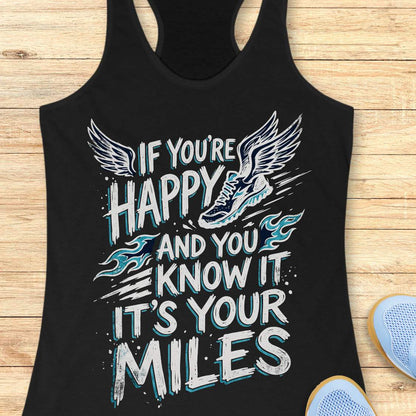 A black racerback tank top laid flat on a light wood background, featuring a large white and light blue graphic with wings, a running shoe, and the text 'IF YOU'RE HAPPY AND YOU KNOW IT IT'S YOUR MILES.' Partial blue shoes are visible at the bottom right as styling props.