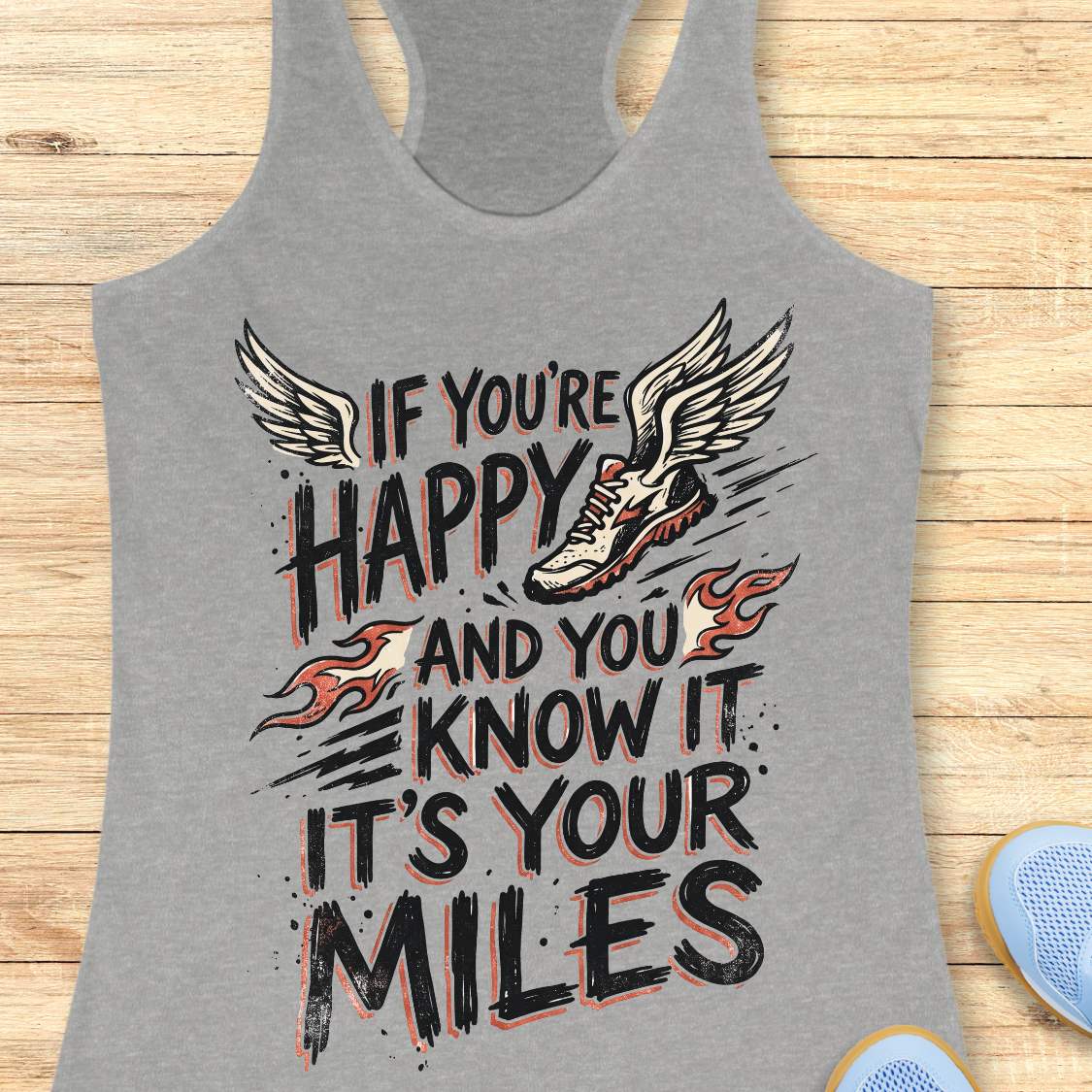 A heather gray racerback tank top laid flat on a light wood background, featuring a large graphic print with the text 'IF YOU'RE HAPPY AND YOU KNOW IT IT'S YOUR MILES' in black, white, and orange-red flame accents, plus a winged running shoe illustration.