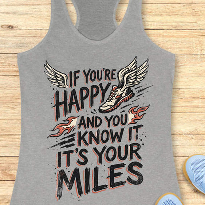 A heather gray racerback tank top laid flat on a light wood background, featuring a large graphic print with the text 'IF YOU'RE HAPPY AND YOU KNOW IT IT'S YOUR MILES' in black, white, and orange-red flame accents, plus a winged running shoe illustration.