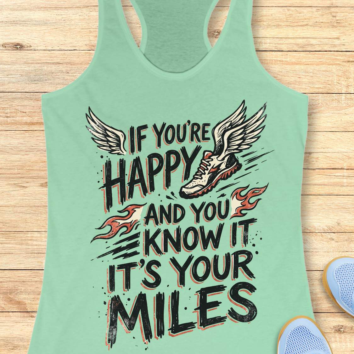 A mint green racerback tank top laid flat on a light wood background, featuring a graphic print with the text 'IF YOU'RE HAPPY AND YOU KNOW IT IT'S YOUR MILES' in black with orange outlines, accompanied by a winged running shoe and small flame motifs.