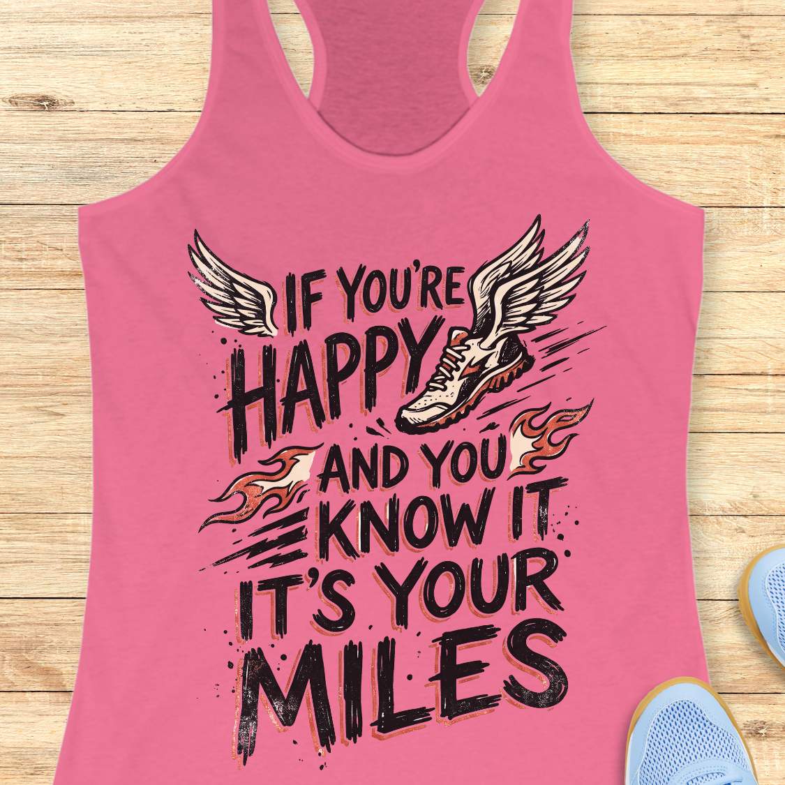 A pink racerback tank top laid flat on a light wood background, featuring a motivational graphic that reads 'IF YOU'RE HAPPY AND YOU KNOW IT IT'S YOUR MILES' with a winged sneaker and flame accents.