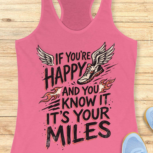 A pink racerback tank top laid flat on a light wood background, featuring a motivational graphic that reads 'IF YOU'RE HAPPY AND YOU KNOW IT IT'S YOUR MILES' with a winged sneaker and flame accents.