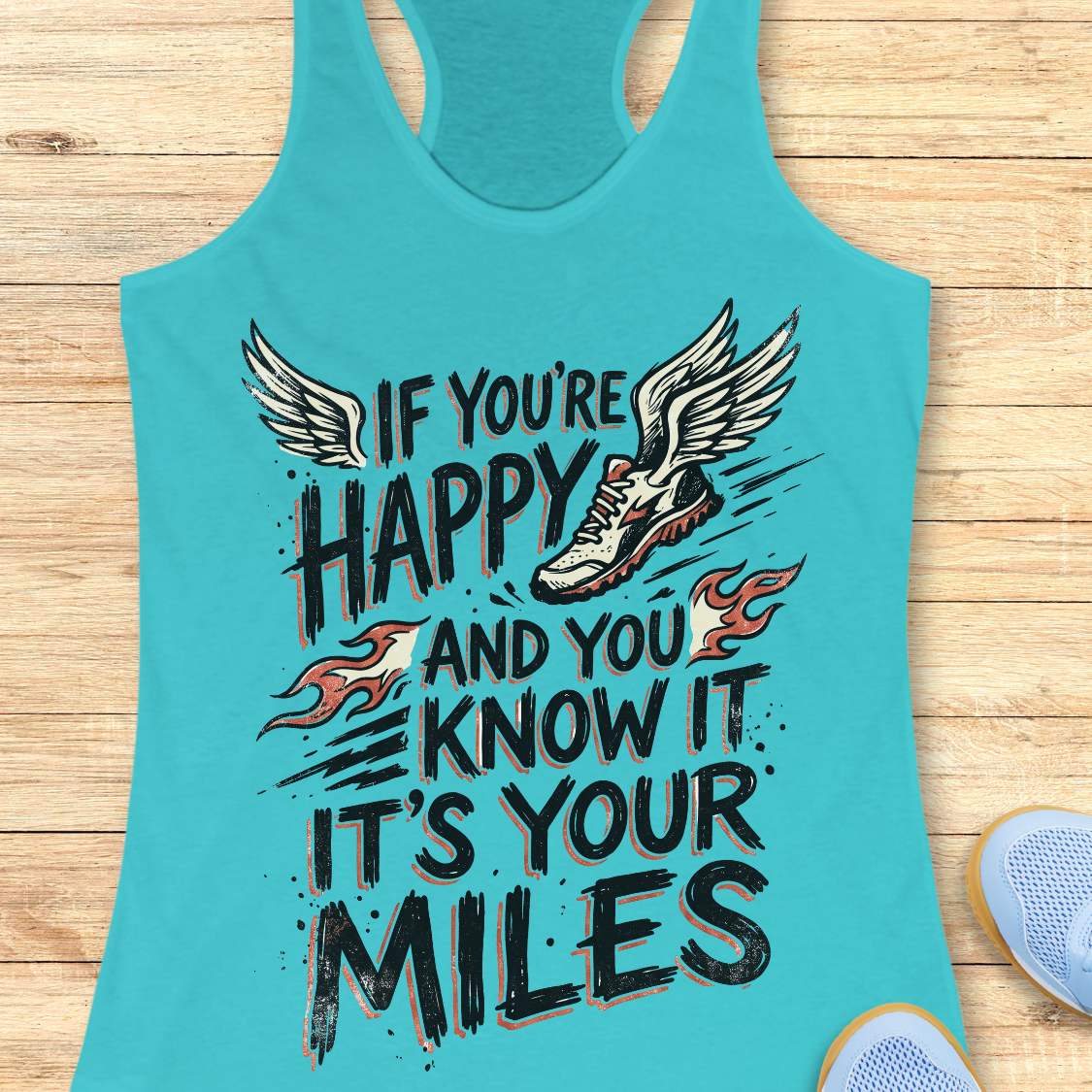 A turquoise racerback tank top laid flat on a light wood surface, featuring a large graphic print with the words 'IF YOU'RE HAPPY AND YOU KNOW IT IT'S YOUR MILES' in black, a winged running shoe, and small flame motifs. Partial blue sneakers are visible at the lower right as styling props.