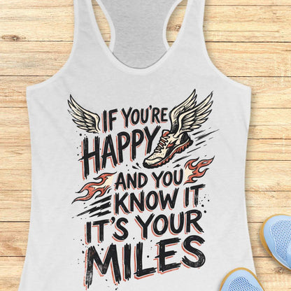 A white racerback tank top laid flat on a light wood background, featuring a graphic print with the text 'IF YOU'RE HAPPY AND YOU KNOW IT IT'S YOUR MILES' in black and red, alongside a winged running shoe and small flame motifs.