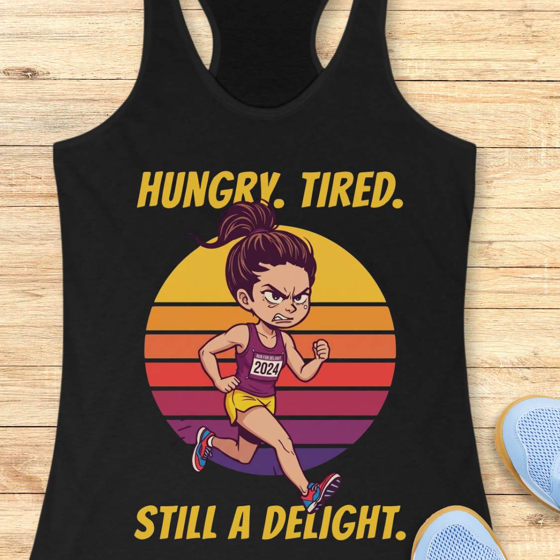A black racerback tank top laid flat on a light wood surface, featuring a colorful graphic of a cartoon runner in front of a retro striped sunset with the text 'HUNGRY. TIRED. Still a delight.'