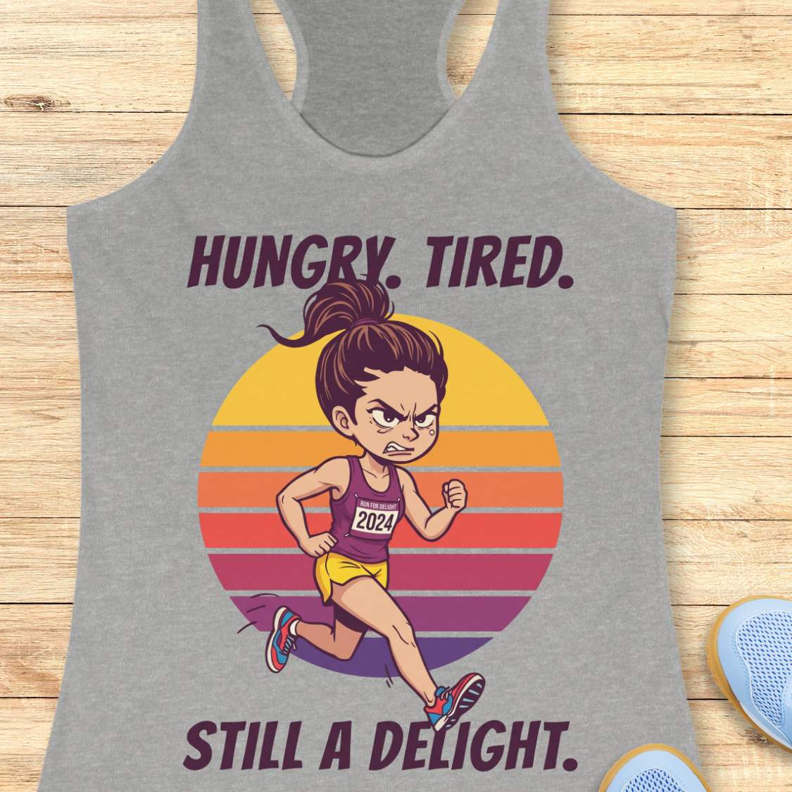 A heather gray racerback tank top laid flat on a light wood surface, featuring a colorful graphic of a cartoon runner in front of a striped sunset with the text 'HUNGRY. TIRED.  Still a delight.'
