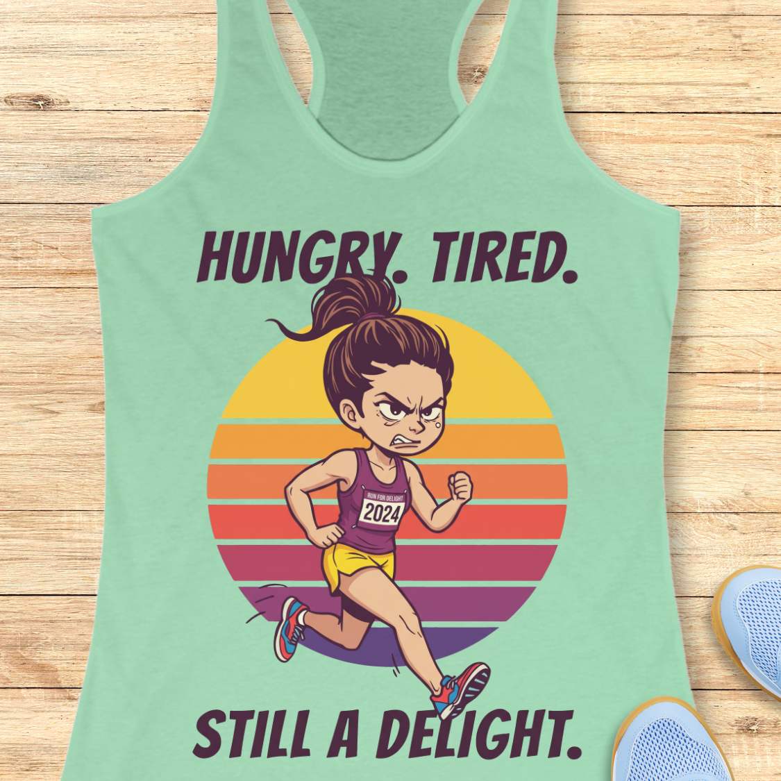 A mint green racerback tank top laid flat on a light wood background, featuring a colorful graphic of a runner in front of a striped sunset with the text 'HUNGRY. TIRED. Still a delight.'