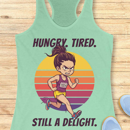A mint green racerback tank top laid flat on a light wood background, featuring a colorful graphic of a runner in front of a striped sunset with the text 'HUNGRY. TIRED. Still a delight.'