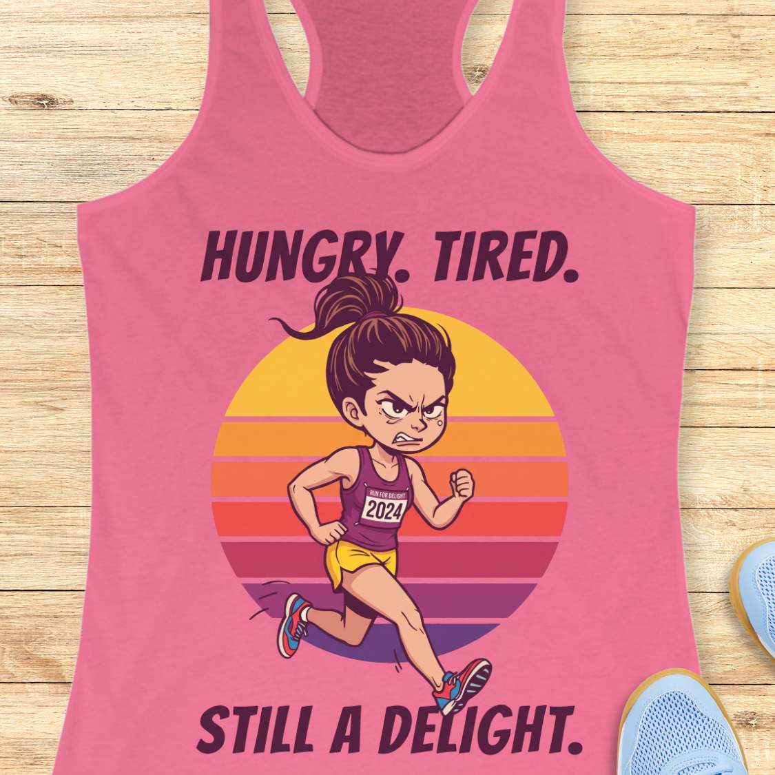 A pink racerback tank top laid flat on a light wood surface, featuring a printed graphic of a cartoon runner in front of a retro striped sunset with the text 'HUNGRY. TIRED.  Still a delight.'