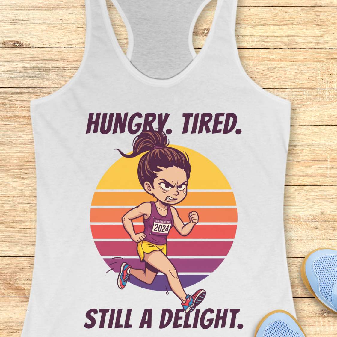 A white racerback tank top laid flat on a light wood surface, featuring a colorful graphic of a cartoon runner in front of a striped sunset with the text 'HUNGRY. TIRED.  Still a delight.'