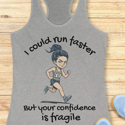 A heather gray racerback tank top laid flat on a light wood background, featuring a cartoon illustration of a female runner with the text 'I could run faster But your confidence is fragile'.
