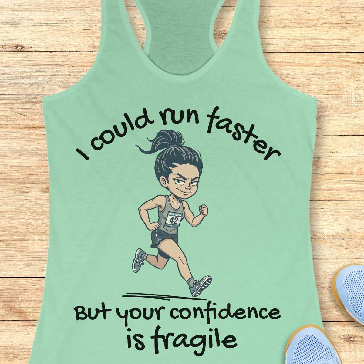 A mint green racerback tank top laid flat on a light wood background, featuring a cartoon illustration of a female runner with the text 'I could run faster But your confidence is fragile'.