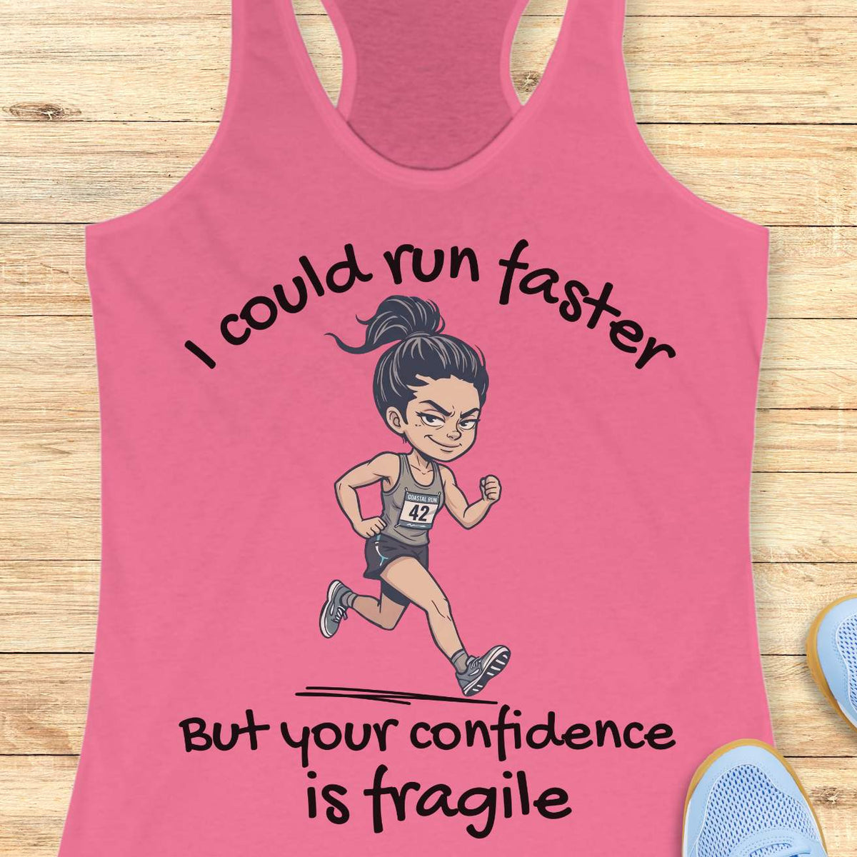 A pink racerback tank top laid flat on a light wood background, featuring a cartoon runner graphic and the text 'I could run faster But your confidence is fragile'.