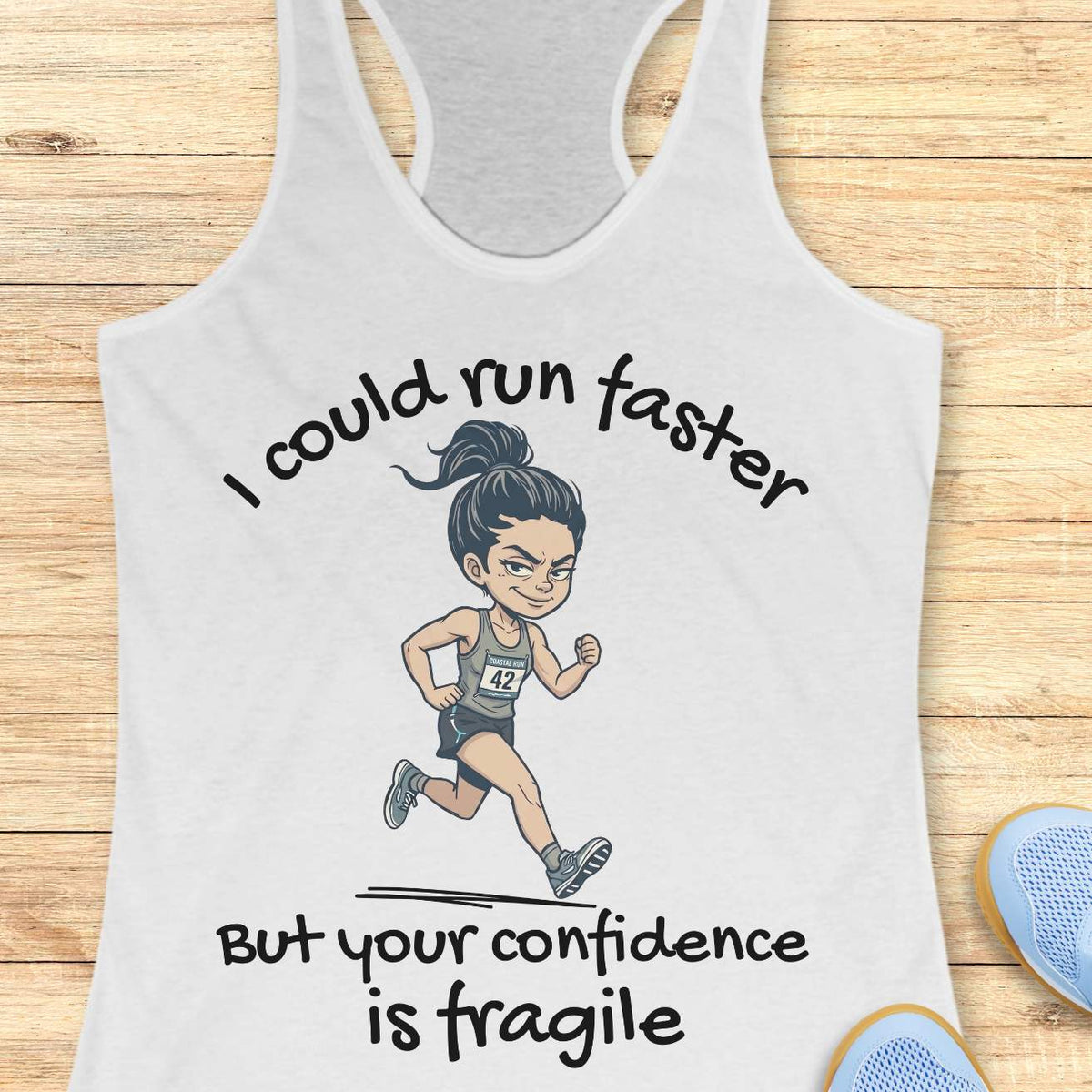 A white racerback tank top laid flat on a light wood background, featuring a cartoon illustration of a female runner and the text 'I could run faster But your confidence is fragile'. Partial blue shoes are visible at the bottom right as styling props.