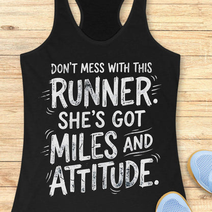 A black racerback tank top with a white distressed text graphic reading 'DON'T MESS WITH THIS RUNNER. SHE'S GOT MILES AND ATTITUDE.