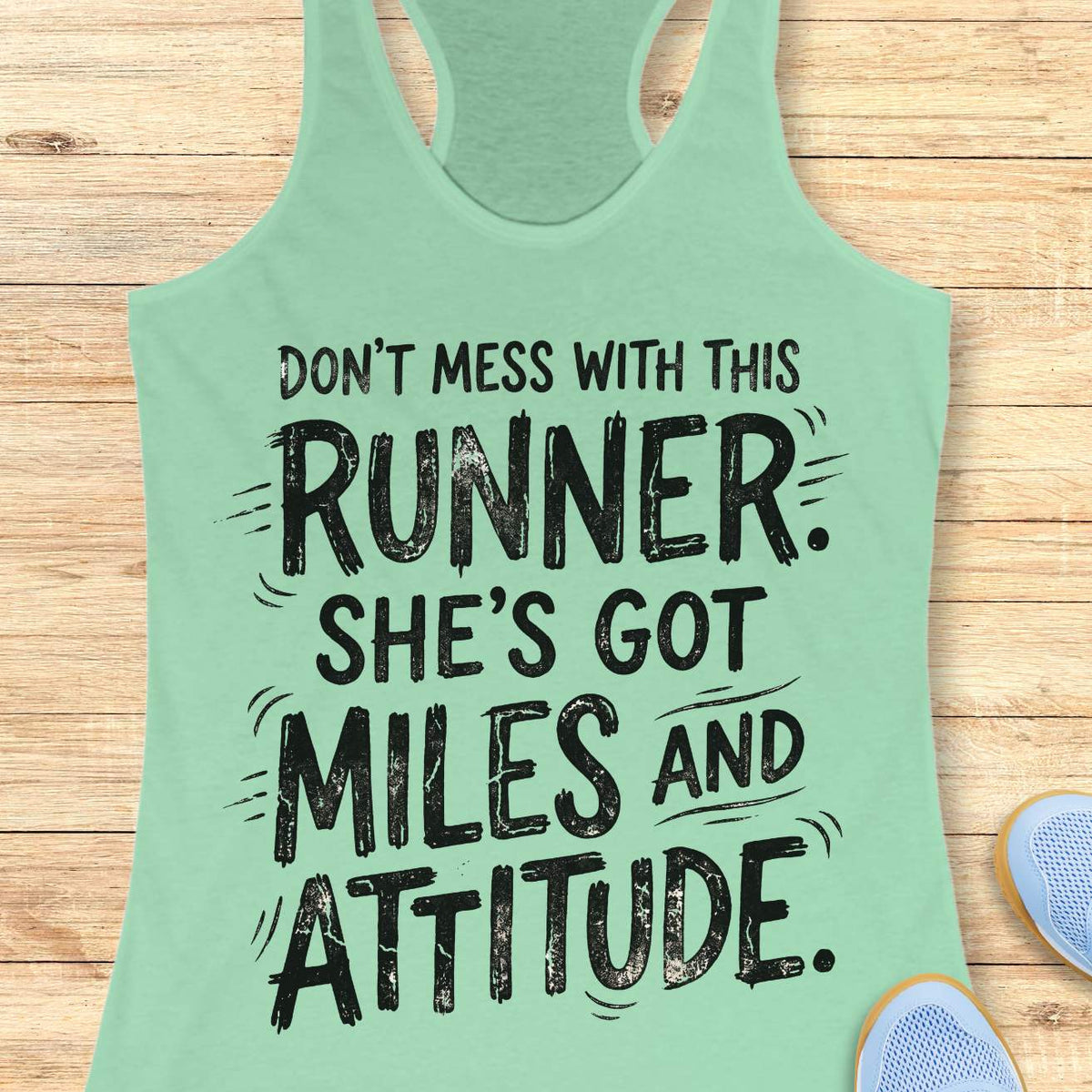 A mint green racerback tank top with a black distressed graphic text reading 'DON'T MESS WITH THIS RUNNER. SHE'S GOT MILES AND ATTITUDE.