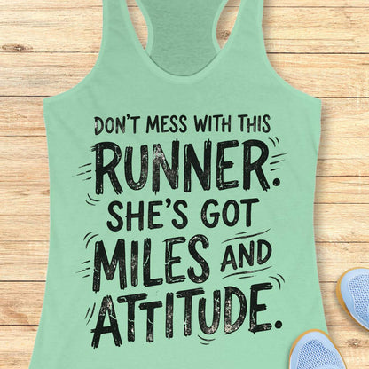 A mint green racerback tank top with a black distressed graphic text reading 'DON'T MESS WITH THIS RUNNER. SHE'S GOT MILES AND ATTITUDE.