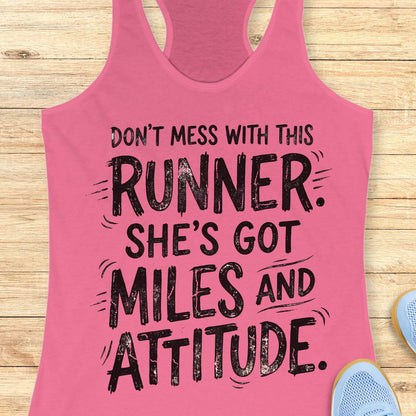 A pink racerback tank top with a black distressed graphic text reading 'DON'T MESS WITH THIS RUNNER. SHE'S GOT MILES AND ATTITUDE.