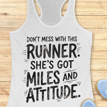A white racerback tank top with a black distressed text graphic reading 'DON'T MESS WITH THIS RUNNER. SHE'S GOT MILES AND ATTITUDE.