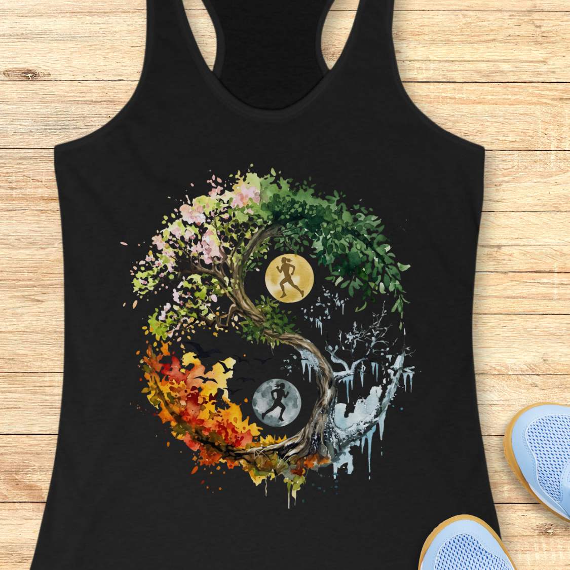 A black racerback tank top laid flat on a light wood surface, featuring a large multicolor Yin Yang graphic with a running figure and autumn leaves. Partial blue sneakers are visible at the bottom right as styling props.