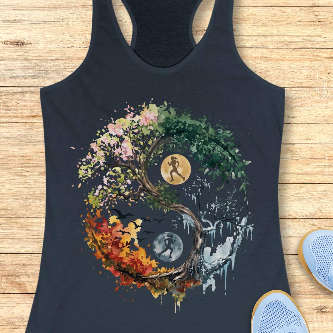 A indigo racerback tank top laid flat on a light wood surface, featuring a large yin-yang style graphic made from a tree with pink, green, orange, and white sections, plus moon and bird motifs. Part of a blue shoe is visible at the lower right edge.