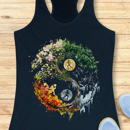 A navy racerback tank top laid flat on a light wood surface, featuring a large circular yin-yang style graphic made from a tree with pink, green, yellow, orange, and white elements, plus running figures and moon icons. Part of a white sneaker is visible at the lower right edge.