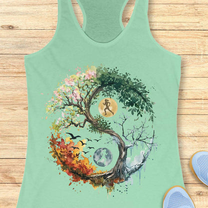 A mint green racerback tank top featuring a large, colorful graphic of a yin-yang tree with seasonal elements and a running figure, displayed flat on a light wood plank background.