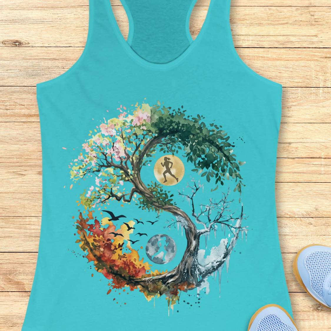 A turquoise racerback tank top laid flat on a light wood surface, featuring a large colorful graphic of a yin-yang tree with autumn leaves, a running figure, birds, and a moon. Part of a blue shoe is visible at the bottom right.